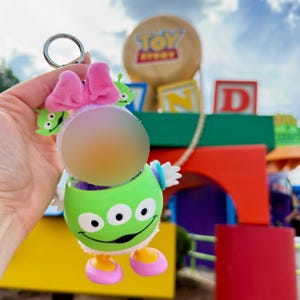 May include: A green alien keychain with a pink bow, white wings, and yellow and pink feet. The keychain has a silver ring and is a fun accessory for fans of the Toy Story franchise.