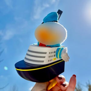 Könnte beinhalten: Eine neuartige Figur eines Zeichentrickcharakters auf einem Kreuzfahrtschiff. Das Schiff ist schwarz und weiß mit gelben Akzenten. Die Figur trägt eine blaue Matrosenmütze und Jacke. Der Hintergrund ist ein strahlend blauer Himmel.