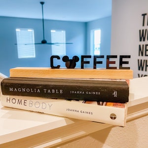 May include: Black acrylic letters spelling "COFFEE" with a Mickey Mouse head in place of the "O". The letters are sitting on top of two books, "Magnolia Table" and "Homebody", both by Joanna Gaines.