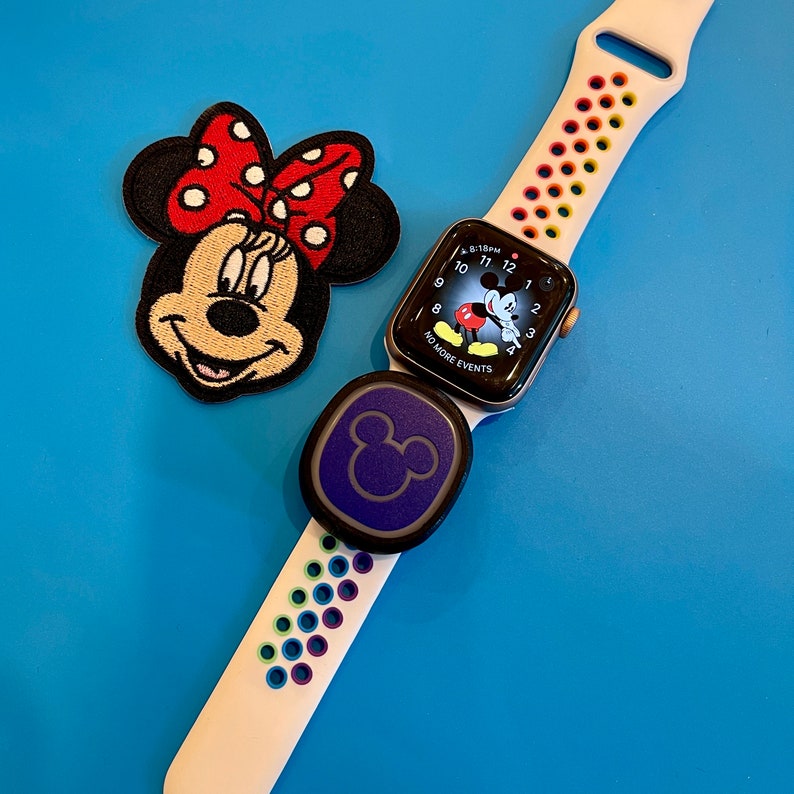 Pu&ograve; includere: Un Apple Watch bianco con un cinturino colorato e un quadrante con il volto di Topolino. L'orologio &egrave; abbinato a un accessorio viola a tema Topolino. L'orologio &egrave; su uno sfondo blu con una toppa di Topolino rossa e nera.