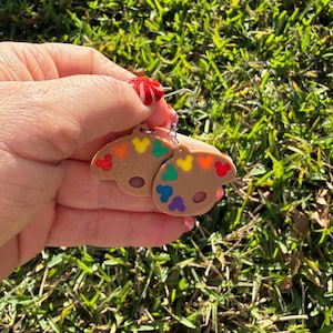 May include: A pair of earrings featuring a brown artist's palette design with colorful paint splotches. The earrings have a silver hook and are held against a backdrop of green grass.