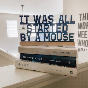 May include: A stack of three books by Joanna Gaines, titled "Magnolia Table", "Magnolia Table", and "Homebody". A black wooden sign with the text "It was all started by a mouse" sits on top of the books.