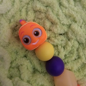 Può includere: Un giocattolo a forma di bruco colorato con una testa arancione, segmenti del corpo gialli, viola e arancione chiaro. Il bruco ha una faccia sorridente ed è ambientato su uno sfondo verde e sfocato.