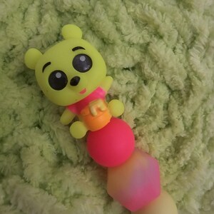 Può includere: Un massaggiagengive per bambini a forma di orso giallo con grandi occhi neri, un bavaglino rosa e un vasetto di miele arancione. Il massaggiagengive ha una fila di perline colorate in rosa, giallo e nero. Lo sfondo è un materiale morbido, verde e sfocato.