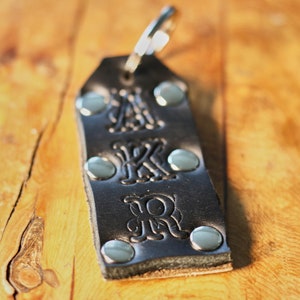 May include: Black leather keychain with a silver metal ring and silver metal rivets. The keychain has the letters "AKR" embossed on it.