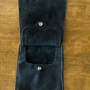 May include: A black leather pouch with a flap and two snap closures. The pouch is open and the inside is visible.