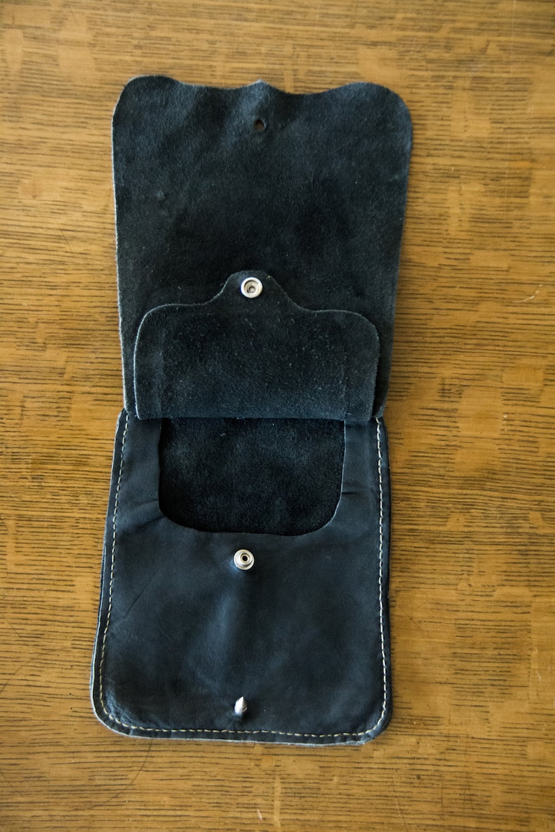 May include: A black leather pouch with a flap and two snap closures. The pouch is open and the inside is visible.