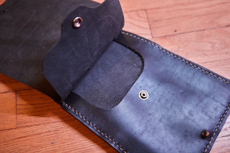 May include: A black leather wallet with a flap that opens to reveal a pocket. The wallet has a stitched edge and three metal snaps.