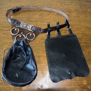 May include: A brown leather belt with a silver buckle and three metal rings. The belt is attached to two black leather pouches. The buckle has the text "Indiana Wildcat" on it.