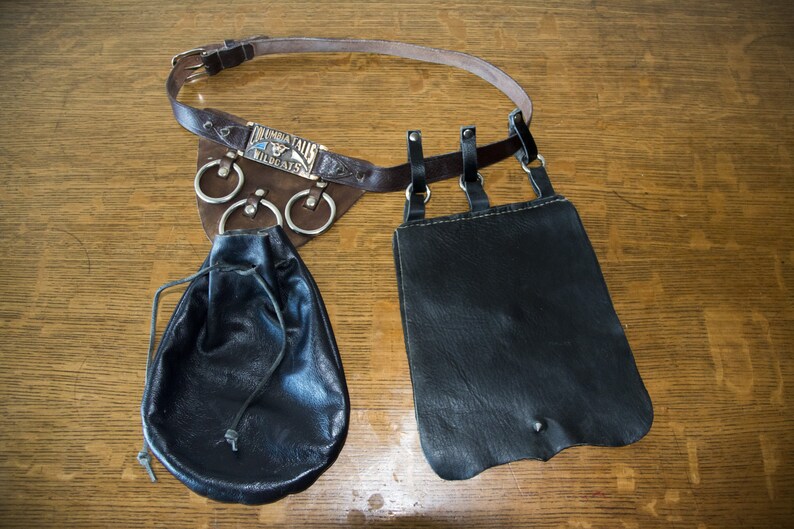 May include: A brown leather belt with a silver buckle and three metal rings. The belt is attached to two black leather pouches. The buckle has the text "Indiana Wildcat" on it.