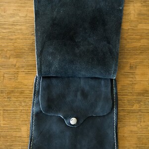 May include: A black leather pouch with a flap and a snap closure. The pouch has a small pocket on the front and is stitched with white thread.