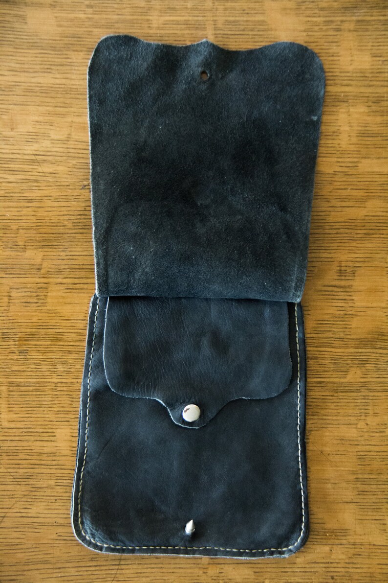 May include: A black leather pouch with a flap and a snap closure. The pouch has a small pocket on the front and is stitched with white thread.