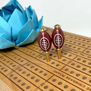 May include: Two brown and white football-shaped cribbage pegs on a wooden cribbage board. The board has a brown wood finish and is marked with numbers and lines.