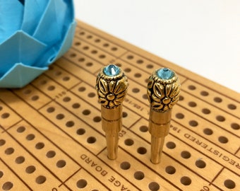 May include: Two gold-colored cribbage pegs with floral designs and light blue gemstones on top. The pegs are on a wooden cribbage board with drilled holes and number markings. A blue origami flower is in the background.