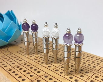 May include: Six silver metal game pieces with different colored tops. Three pieces have purple amethyst tops and three pieces have white sparkly tops. The pieces are on a wooden game board with numbered holes.