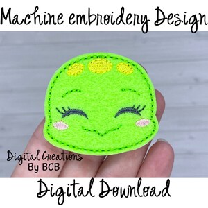 May include: A green felt applique of a smiling dinosaur with yellow spots and pink cheeks. The dinosaur is being held in a hand. The text "Digital Creations By BCB" and "Digital Download" are visible below the dinosaur.