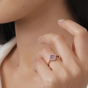 May include: A rose gold ring featuring a square-shaped stone set within a diamond-shaped frame. The stone is a pale purple colour.