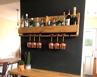Pode incluir: Uma prateleira de bar de parede de madeira com fundo preto. A prateleira tem uma prateleira superior para garrafas e uma prateleira inferior para copos. Há várias garrafas de licor na prateleira superior e uma fileira de copos de vinho de cor cobre pendurados na prateleira inferior. Uma mesa de madeira com pernas de grampo de cabelo pretas fica na frente da prateleira.