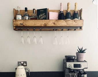 Pode incluir: Um suporte de vinho de madeira rústico com garrafas de licor e champanhe, livros e taças. Seis taças de vinho penduradas embaixo. Uma chaleira branca e uma máquina de café expresso estão na bancada.