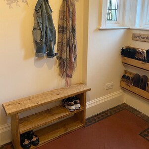 May include: A wooden hallway bench with two shelves for shoes. The bench has a coat rack above it with hooks and a shelf for decorative items. The wall is white and there is a sign that says "Family" above the coat rack. There is a small wooden shelf with shoes on it in the corner of the room.