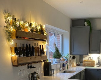 Pode incluir: Uma cozinha moderna com uma bancada branca e uma ilha azul com dois banquetas de bar cinzas. A ilha tem uma prateleira de vinho integrada com uma corda de luzes e uma pequena planta em vaso. A cozinha tem armários cinzas e um exaustor de aço inoxidável.