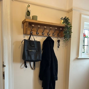 May include: A wooden coat rack with a shelf and hooks, a black backpack hanging on one hook, a black coat hanging on another hook, and a set of keys hanging on a hook. A wooden bench with two shelves is below the coat rack, with shoes on the shelves.