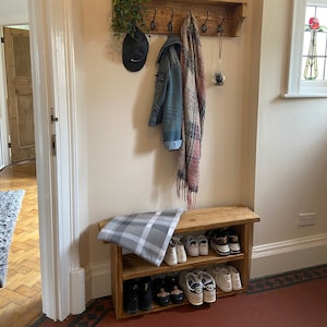 May include: A wooden coat rack with hooks and a shelf, featuring a plant, a hat, a jacket, and a scarf. Below the rack is a wooden shoe rack with two shelves, holding a variety of shoes. The wall is painted a light cream colour. The text on the sign reads "Family is not an important thing. It's everything."