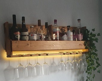 Pode incluir: Um suporte de vinho de madeira montado na parede com uma tira de luz embaixo. O suporte tem uma prateleira para garrafas e um suporte inferior para pendurar copos de vinho. Há várias garrafas de licor na prateleira, incluindo Baileys, Captain Morgan e uma garrafa de gin. Há uma planta verde pendurada do lado direito do suporte.
