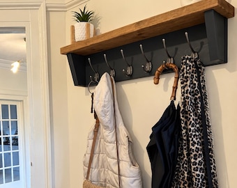 May include: A wall-mounted coat rack with a natural wood shelf and a black backboard featuring seven silver metal hooks. Items hanging include a cream puffer vest, a tan woven crossbody bag, a black umbrella, a leopard print scarf, and a beige bag.