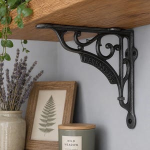 Puede incluir: Un soporte de estante de metal gris oscuro con un diseño de volutas ornamentales y la palabra DERBYSHIRE grabada a lo largo del brazo de soporte diagonal. El soporte está fijado a un estante de madera y a una pared, con orificios para tornillos.