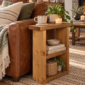 May include: A narrow, rectangular wooden side table with a natural finish, featuring a flat top surface and one middle shelf. The table holds a speckled mug, a small potted plant, a candle, a stone bowl, and two books titled The Kinfolk Home and At Home.