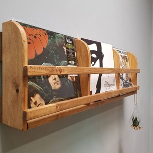 Puede incluir: Una estanter&iacute;a de pared de madera con cuatro discos de vinilo expuestos. La estanter&iacute;a est&aacute; hecha de madera natural con un acabado r&uacute;stico. Los discos incluyen "Rubber Soul" de The Beatles, "The Beatles" de The Beatles, "The Contours" de The Contours y "Stone Roses" de The Stone Roses.