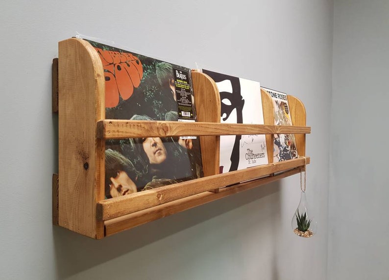 Puede incluir: Una estanter&iacute;a de pared de madera con cuatro discos de vinilo expuestos. La estanter&iacute;a est&aacute; hecha de madera natural con un acabado r&uacute;stico. Los discos incluyen "Rubber Soul" de The Beatles, "The Beatles" de The Beatles, "The Contours" de The Contours y "Stone Roses" de The Stone Roses.