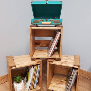 May include: Three wooden crates stacked on top of each other, with a teal record player on top. The crates are filled with vinyl records. The top crate has a small plant on the bottom shelf.
