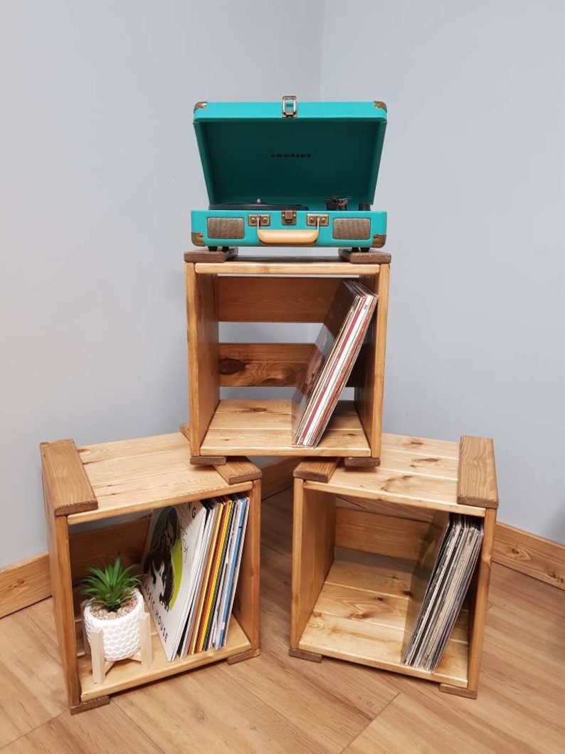 May include: Three wooden crates stacked on top of each other, with a teal record player on top. The crates are filled with vinyl records. The top crate has a small plant on the bottom shelf.