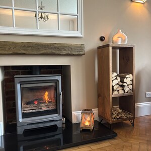 May include: A rustic wooden log holder with three shelves, featuring a white ceramic vase and a candle lantern on the top shelf. The log holder is in front of a fireplace with a black hearth and a burning fire.
