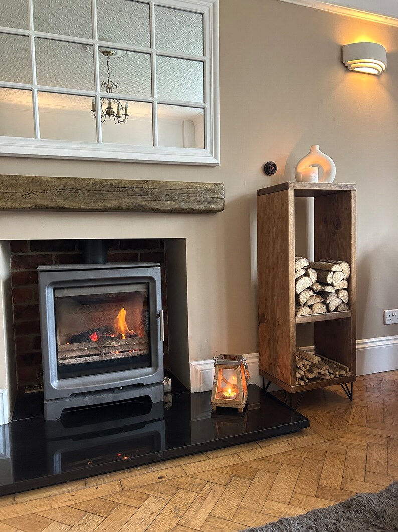 May include: A rustic wooden log holder with three shelves, featuring a white ceramic vase and a candle lantern on the top shelf. The log holder is in front of a fireplace with a black hearth and a burning fire.