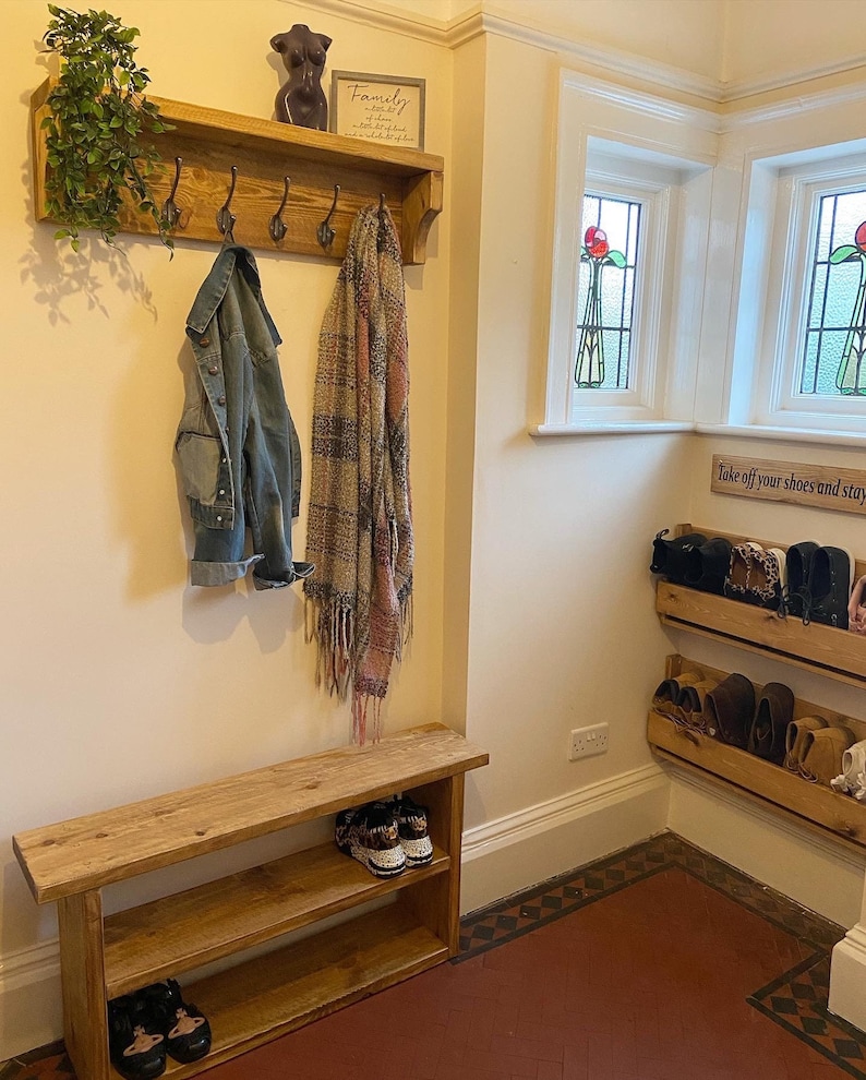 May include: A wooden hallway bench with two shelves for shoes. The bench has a coat rack above it with hooks and a shelf for decorative items. The wall is white and there is a sign that says "Family" above the coat rack. There is a small wooden shelf with shoes on it in the corner of the room.