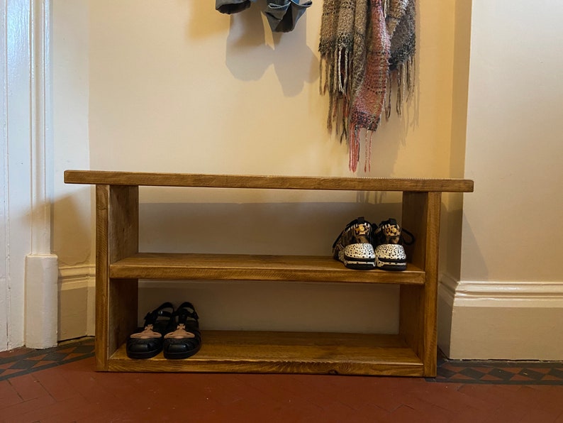 May include: A wooden shoe rack with two shelves and a bench top. The rack is stained a dark brown colour and has a rustic look. There are shoes on the shelves and a scarf hanging on the wall above the rack.