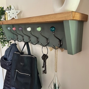 May include: A rustic wooden shelf with a green painted back and a wooden top. The shelf has six hooks with ceramic knobs in various colours. A black backpack, a jacket, and a glass vase with a small plant are hanging on the hooks.