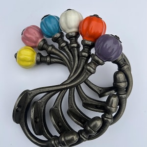 May include: Cabinet hardware with colourful ceramic knobs in pink, blue, white, orange, purple, and yellow. The knobs are mounted on a dark grey metal base with a swirling design. Suitable for drawers and cabinets. Measurements in cm.