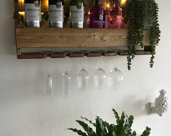 May include: A wooden wall-mounted wine rack with three bottles of red wine and two bottles of gin. The rack has a shelf for bottles and a lower rack for hanging wine glasses. The rack is decorated with string lights and greenery.