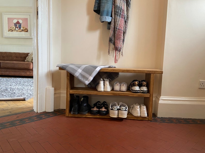 May include: A wooden shoe rack with two shelves, holding a variety of footwear, including trainers, boots, and sandals. The rack is in a hallway with a white door and a brown tiled floor.