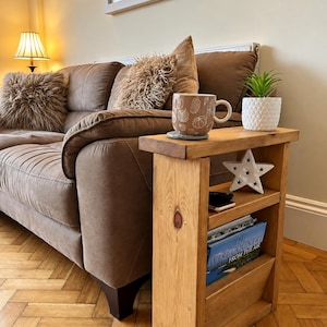 May include: A narrow, rectangular wooden side table with a light brown finish, featuring a top surface, a middle shelf, and a bottom storage compartment. The table holds a brown mug with white pumpkin illustrations, a white star, and a book.