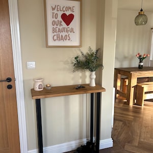 May include: A wooden console table with black metal legs. A framed print with the text "WELCOME TO OUR BEAUTIFUL CHAOS" hangs above the table. A white vase with greenery, a small candle holder, and a pair of black shoes are on the table.