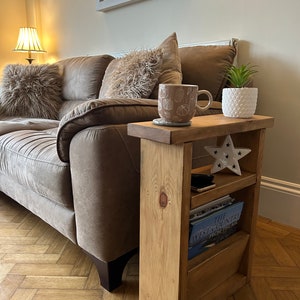 May include: A wooden side table with a shelf and a magazine rack. The table is stained a light brown colour and has a natural wood finish. A mug, a potted plant, and a book are on the table.