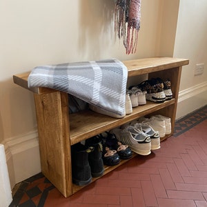 May include: A wooden shoe rack with two shelves, holding a variety of shoes, including trainers, sandals, and boots. The rack is made of natural wood and has a rustic finish. A blanket is placed on top of the rack.