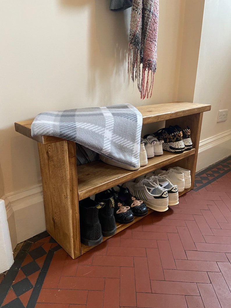 May include: A wooden shoe rack with two shelves, holding a variety of shoes, including trainers, sandals, and boots. The rack is made of natural wood and has a rustic finish. A blanket is placed on top of the rack.