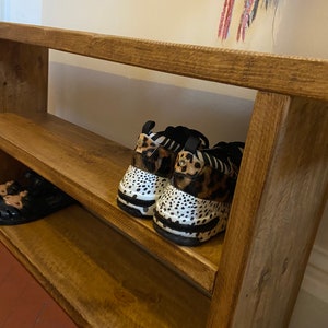 May include: A wooden shoe rack with two shelves. The top shelf has a pair of black and white animal print trainers. The bottom shelf has a pair of sandals.