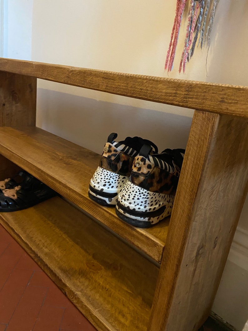 May include: A wooden shoe rack with two shelves. The top shelf has a pair of black and white animal print trainers. The bottom shelf has a pair of sandals.
